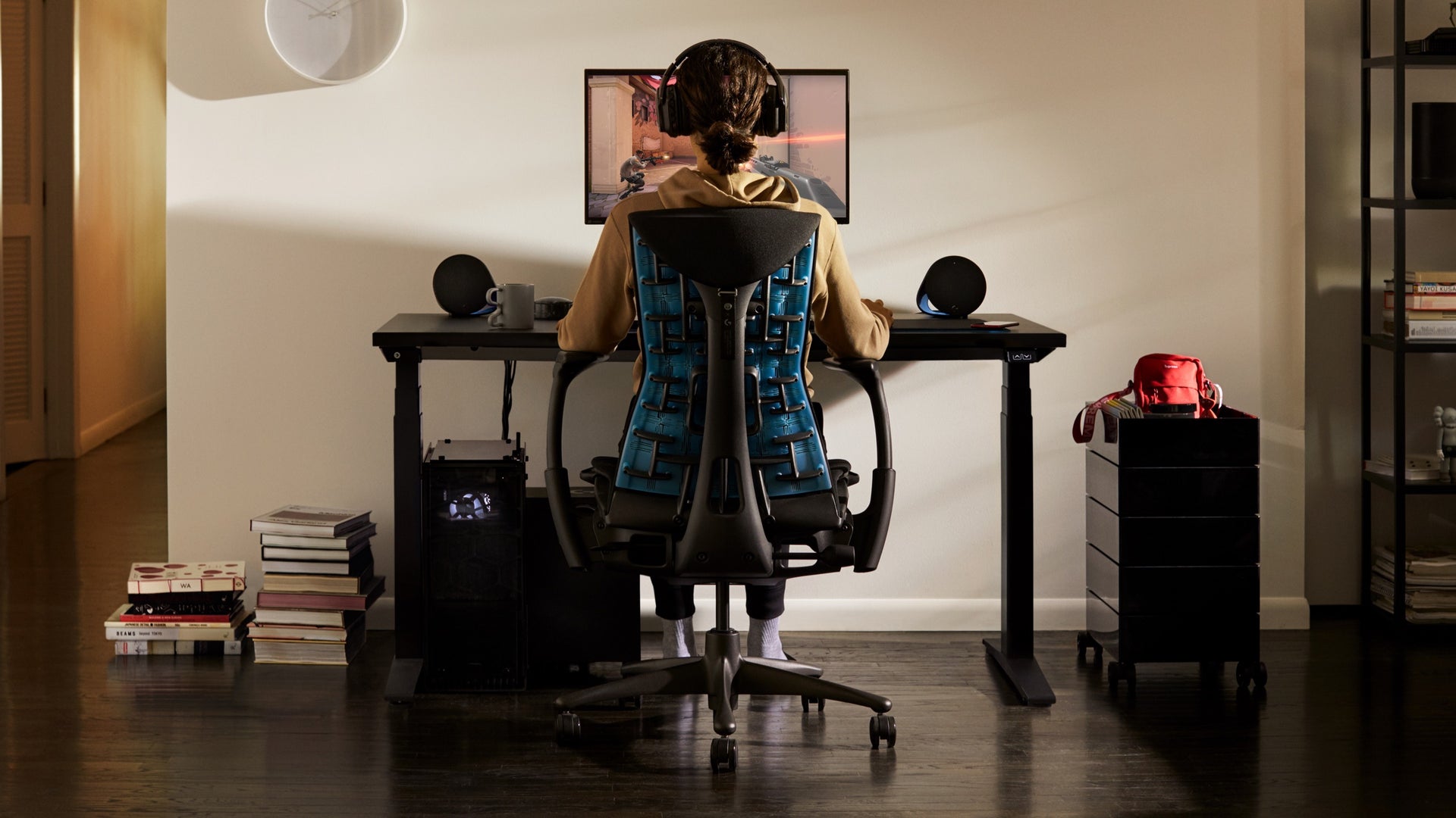 어떤 사람이 Embody Gaming Chair에 앉아서, 주거 환경에서 게이밍 데스트 위에 놓여있는 Ollin Monitor Arm에 부착된 모니터를 봅니다.